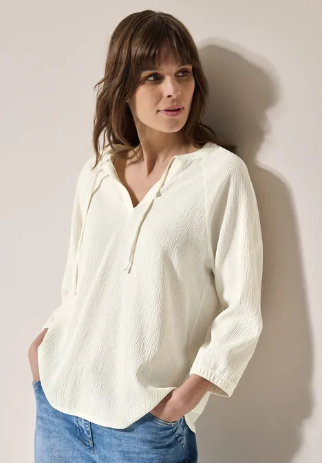 Frau in weißer Bluse mit V-Ausschnitt und Jeans steht vor heller Wand.